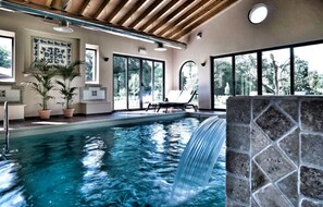 Una piscina con cascada, ventanas amplias y una silla de descanso.