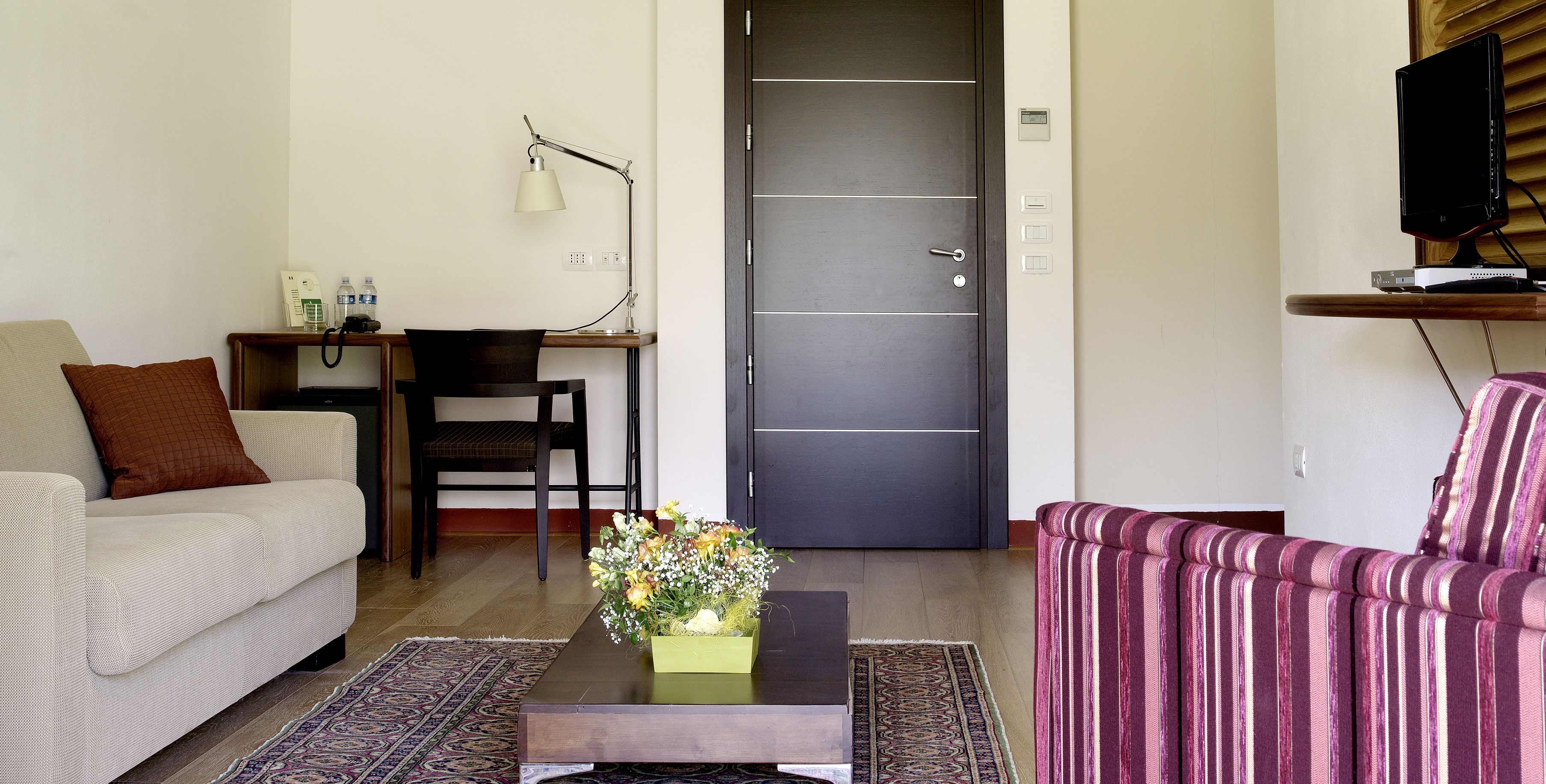 Un salon comprenant un canapé, un fauteuil, une table basse agrémentée d’un arrangement floral, un bureau avec une lampe et un téléviseur posé sur un meuble.