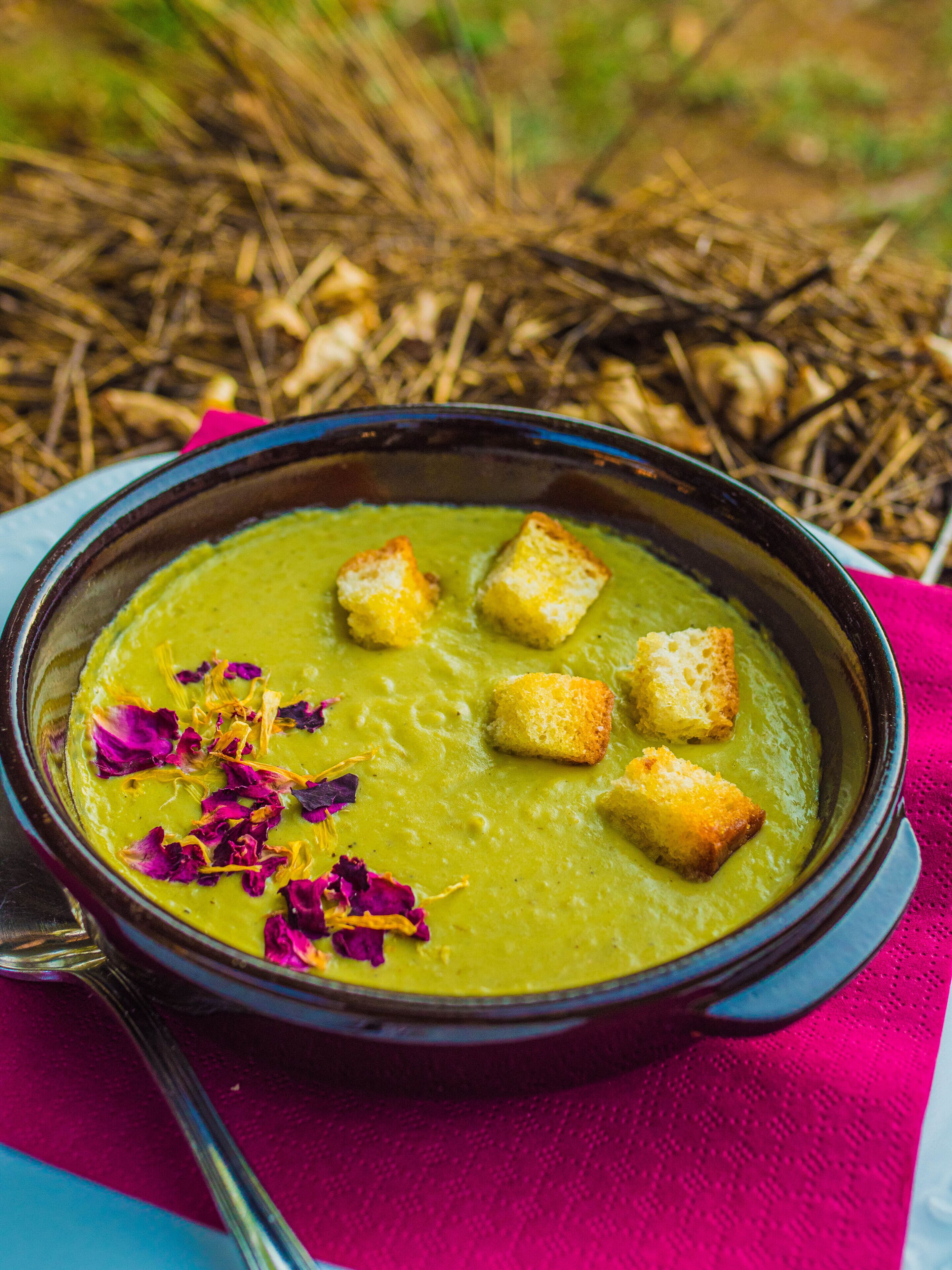 Un tazón de sopa verde con crutones y pétalos de flores, sobre un paño colorido.