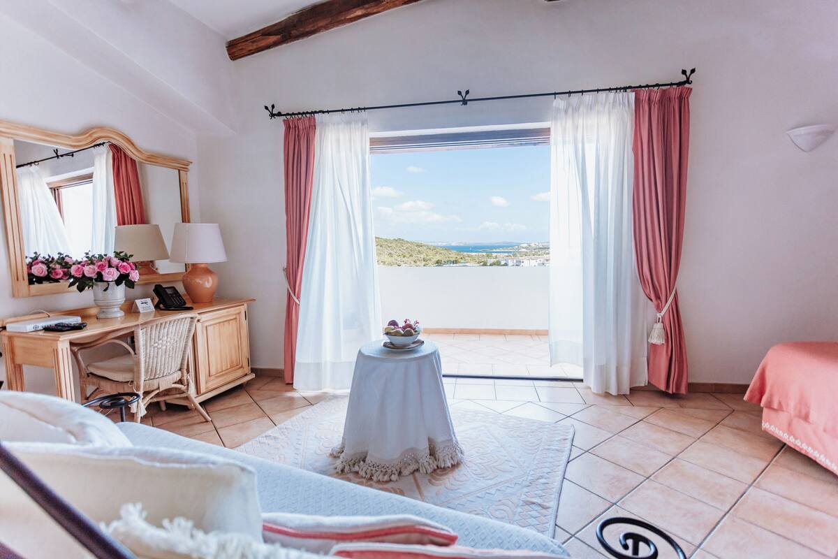 A room with a bed, a small table with a lamp, a dresser, and a view of the sea.