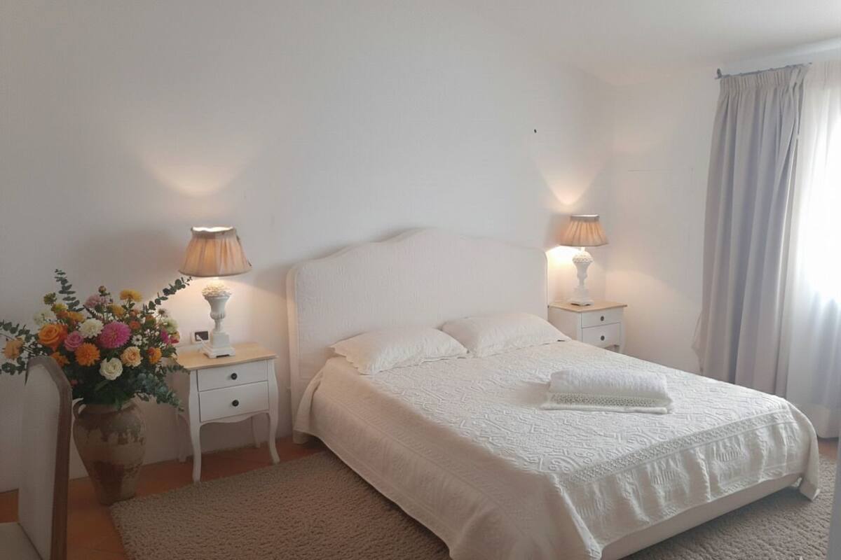 A bedroom with a large bed, two bedside tables with lamps, a vase of flowers, and a window with curtains.