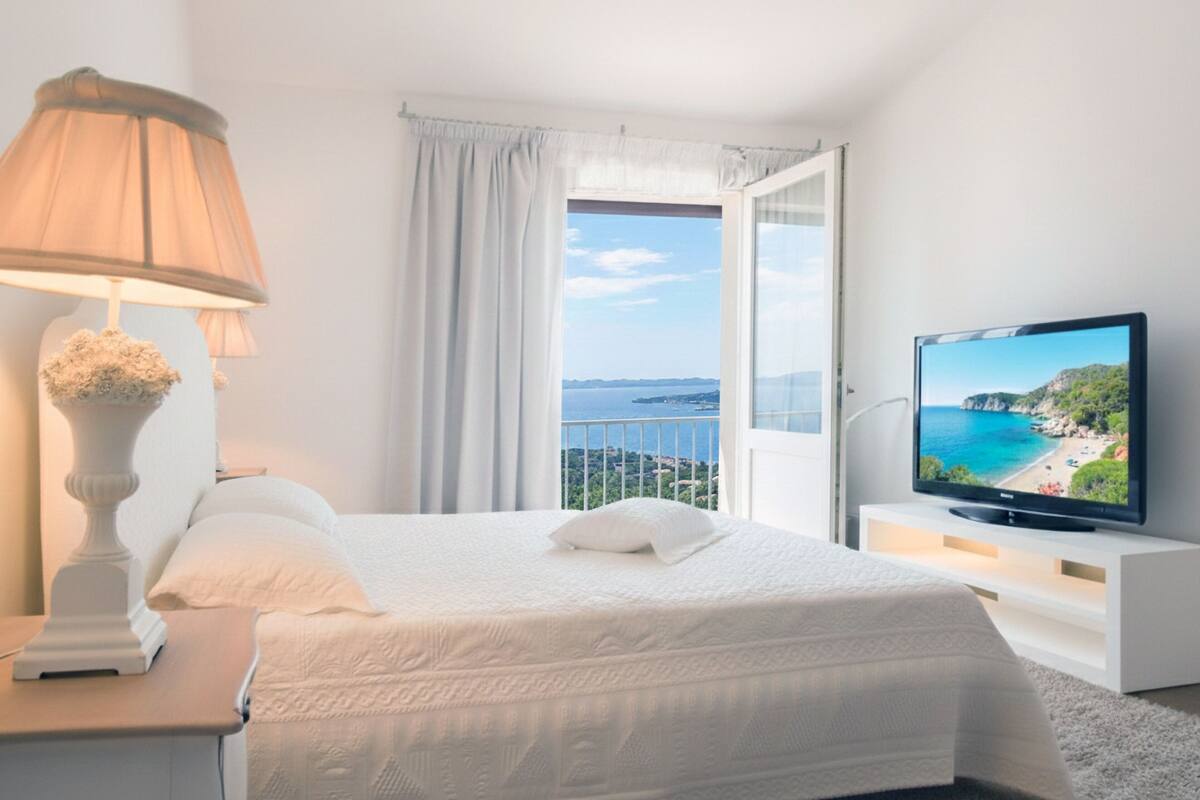 A bedroom with a bed, a TV, a lamp, and a view of the sea.