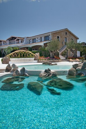 A pool with clear water and rocks, a white building with a tiled roof, and a bridge in the background. - Hotel Pulicinu (Arzachena)