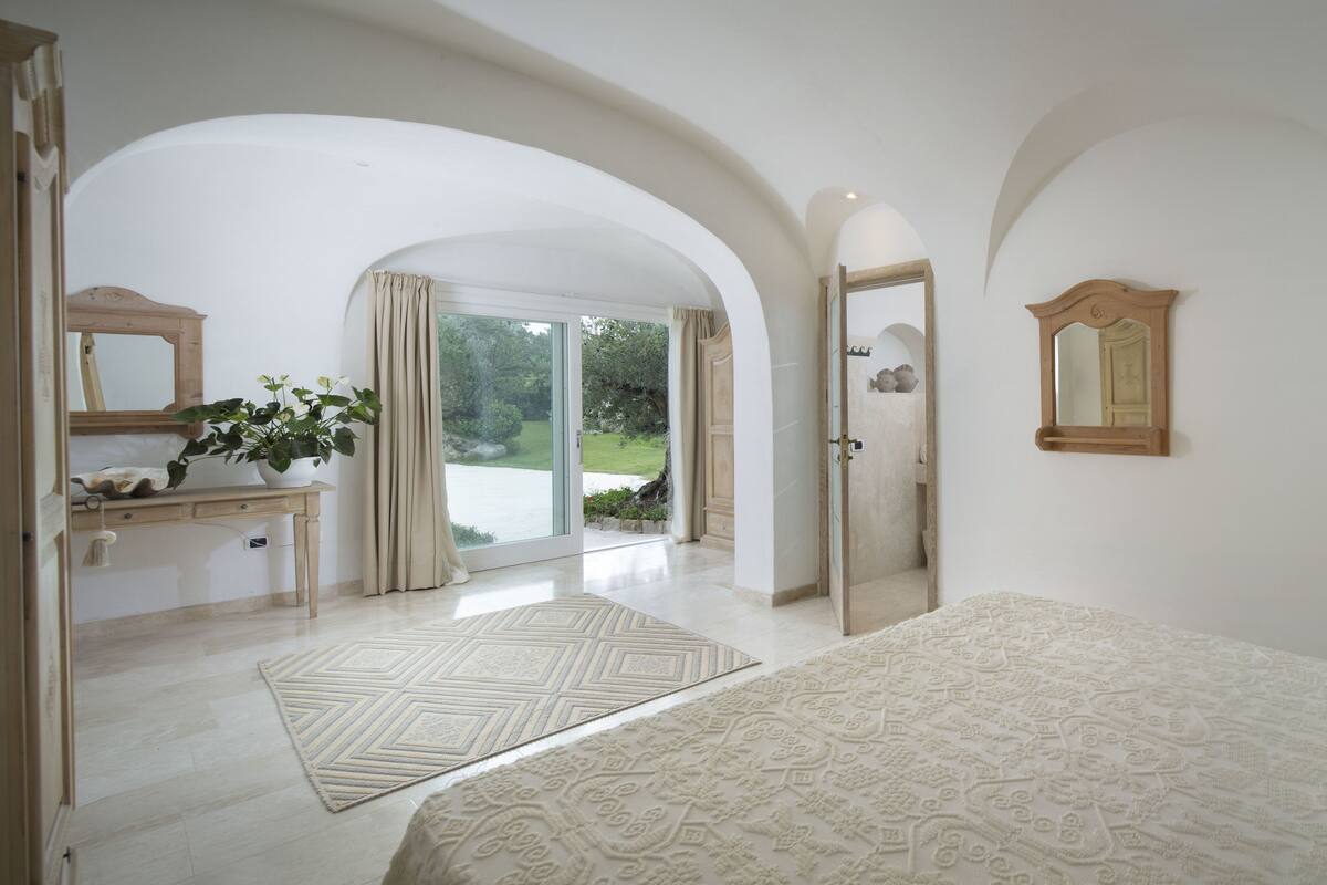 A bedroom with a bed, a nightstand, a lamp, a mirror, and a view of a garden through an open door.