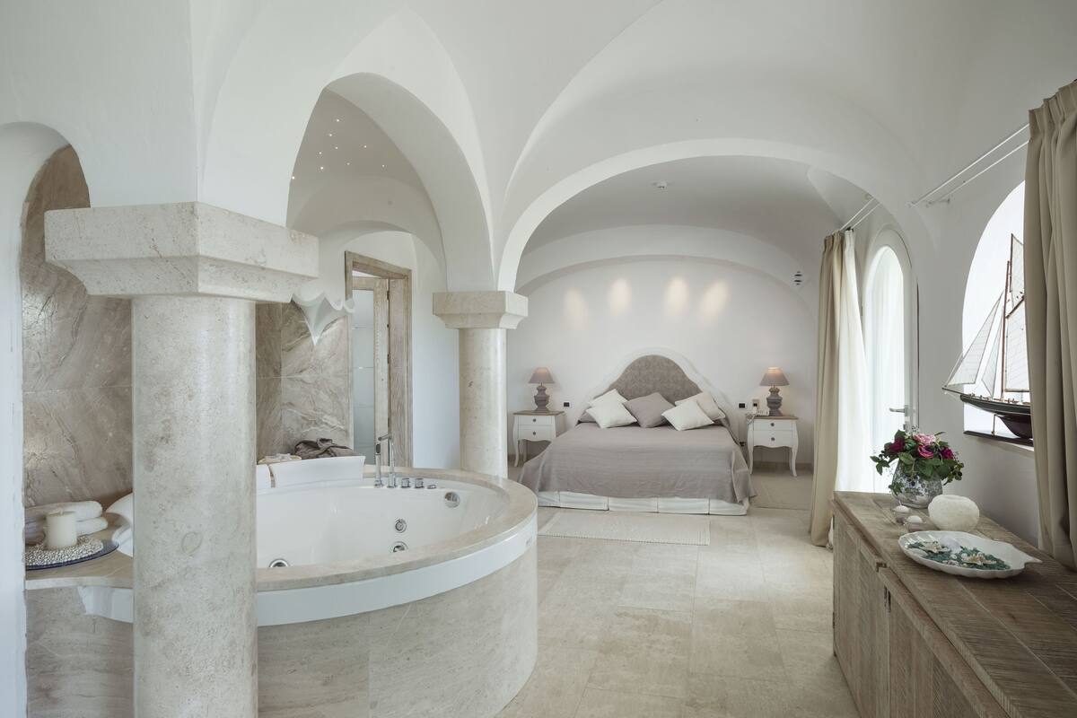 A spacious bedroom with a large curved bathtub, a bed with a headboard, and a view of the sea through a window.
