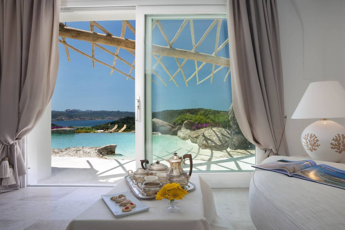 A room with a view of a beach and ocean, a table with tea set and cookies, and a lamp on a bedside table.