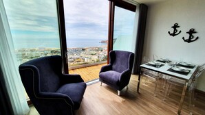 Uma varanda com duas poltronas azul-escuras, uma mesa de jantar posta com pratos e copos, e vista para o horizonte costeiro.