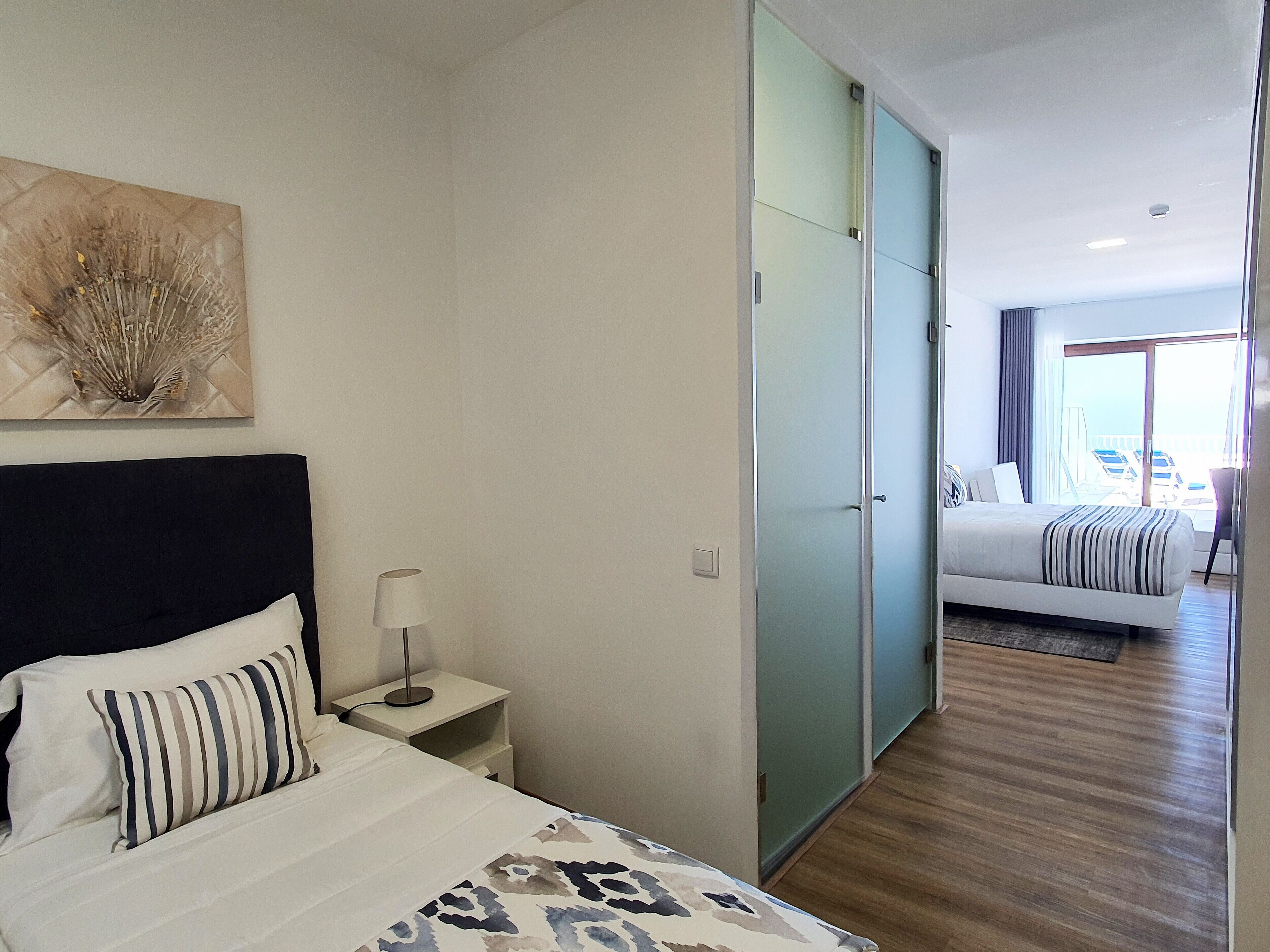 A hotel room with a bed, a nightstand, a lamp, a wall art piece, and a sliding door leading to another room with a balcony.