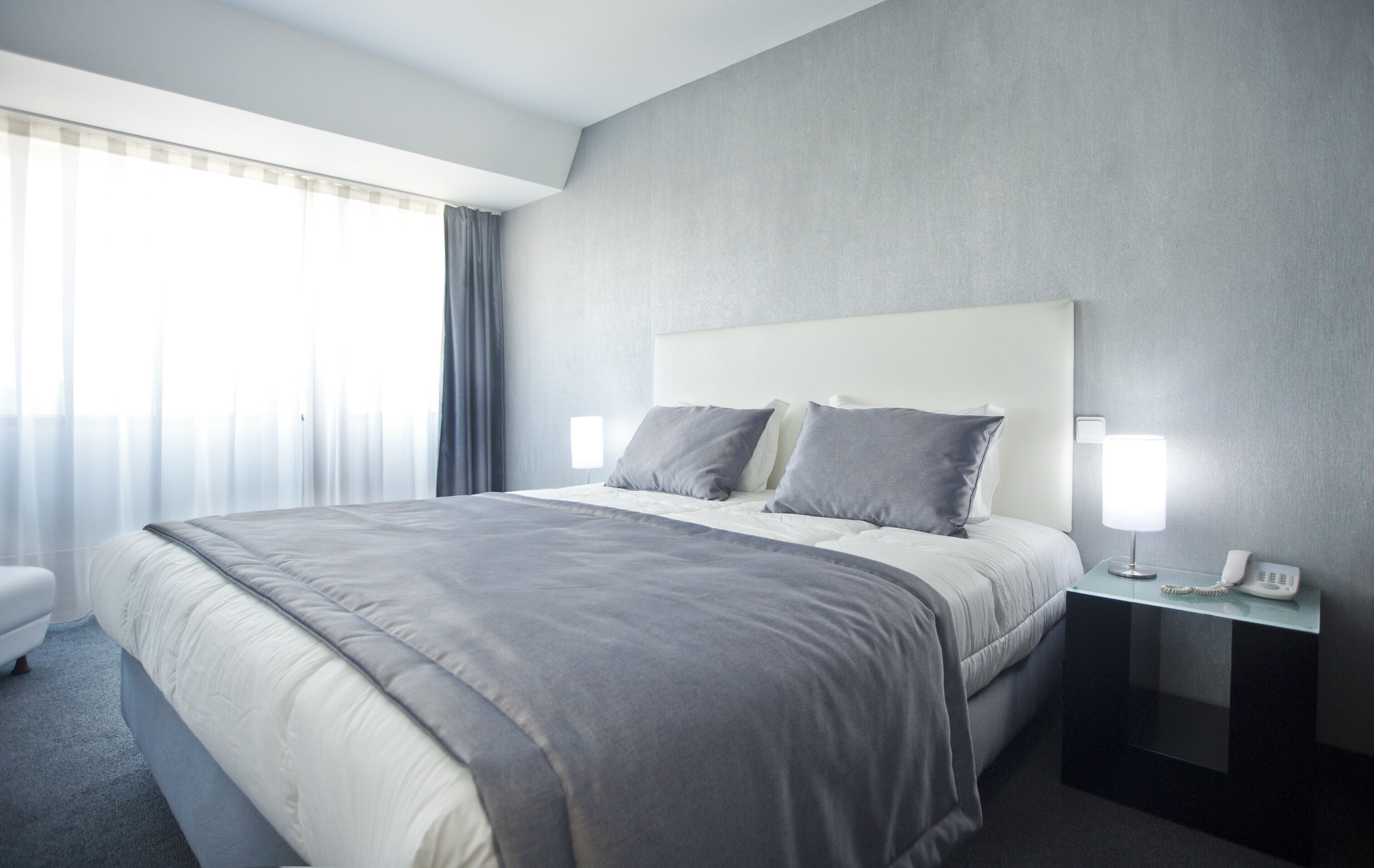 A modern hotel room with a large bed, grey bedding, a bedside table with a lamp, and a phone.
