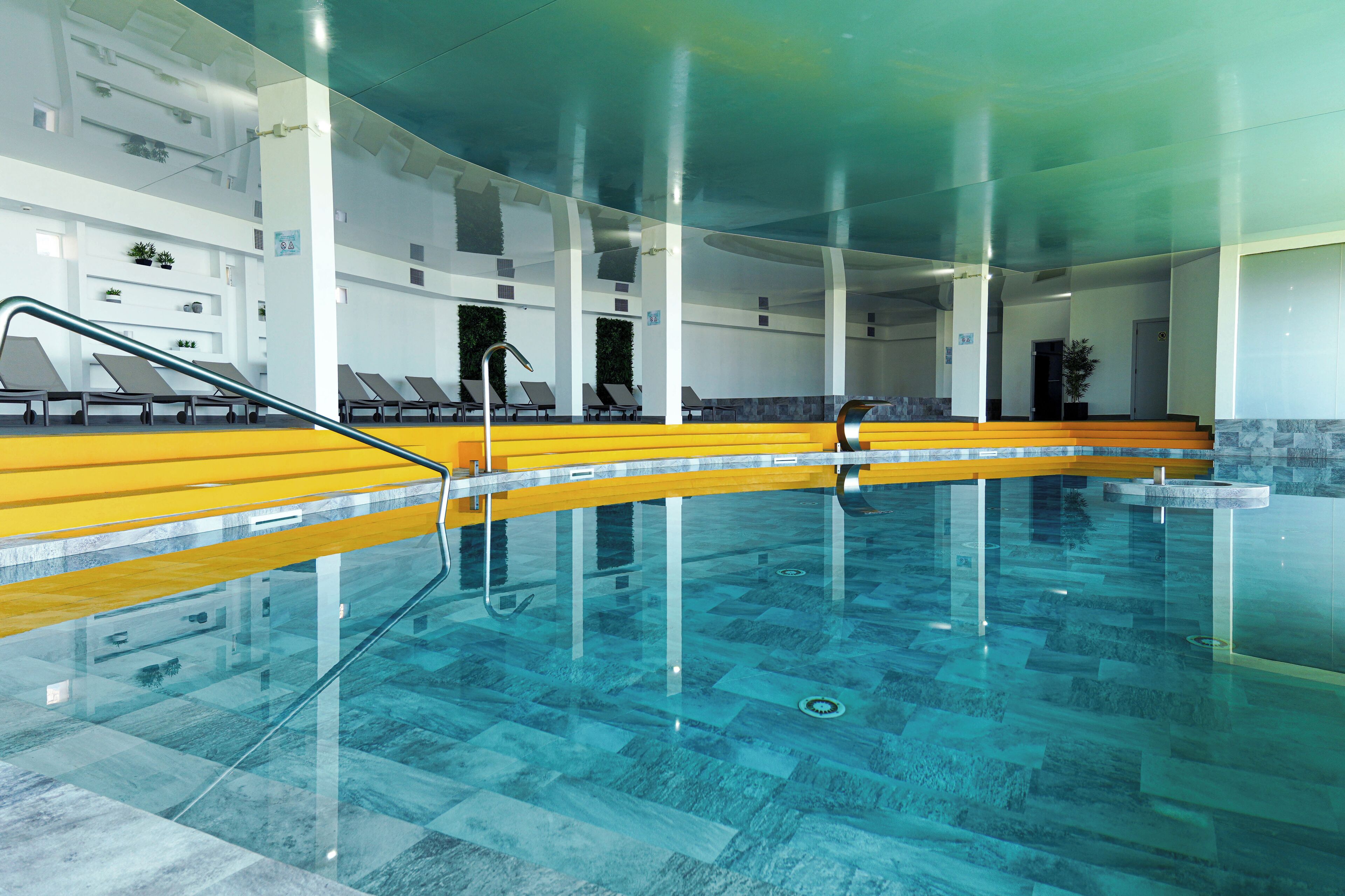 An indoor swimming pool with clear glass walls, yellow steps, and lounge chairs.