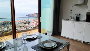 Uma área de refeições com uma mesa posta para dois, vista para o mar e uma vila costeira, além de uma cozinha compacta com armários e pia.