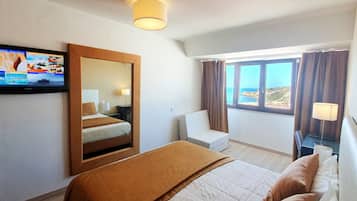 A hotel room with a bed, a TV mounted on the wall, a window with curtains, and a view of the sea.