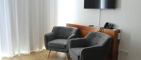 A hotel room with two grey armchairs, a wooden desk with a flat-screen TV, and a white wall.