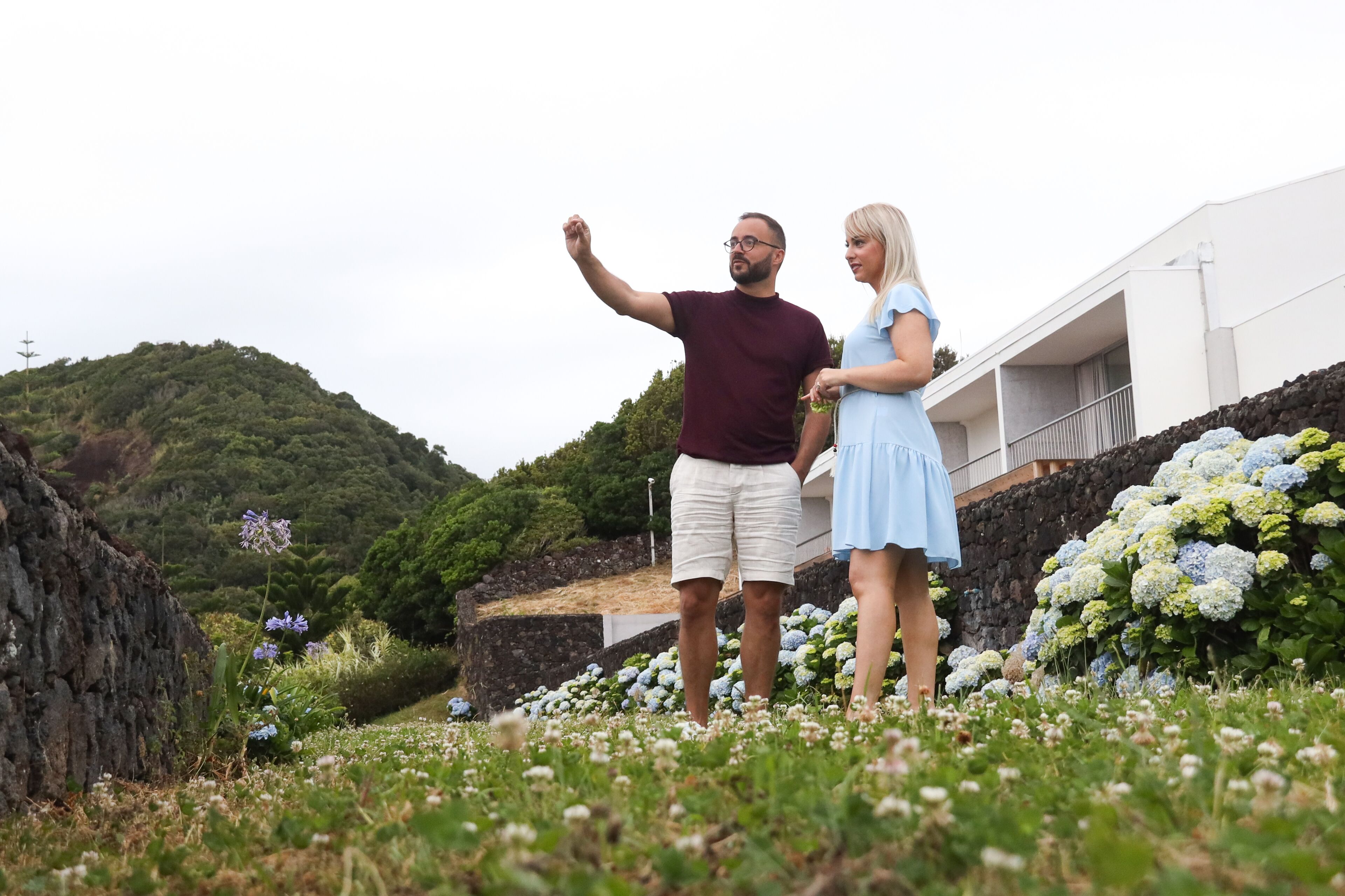 A couple standing in a garden with a view of a hillside and a building in the background.