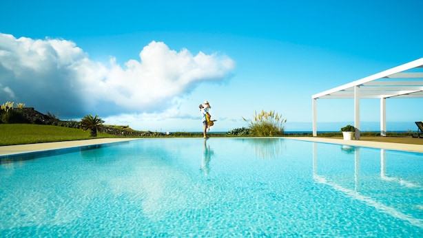 A clear blue swimming pool with a person standing in the center, surrounded by greenery and a white structure on the right. - ANC Resort (Lagoa)