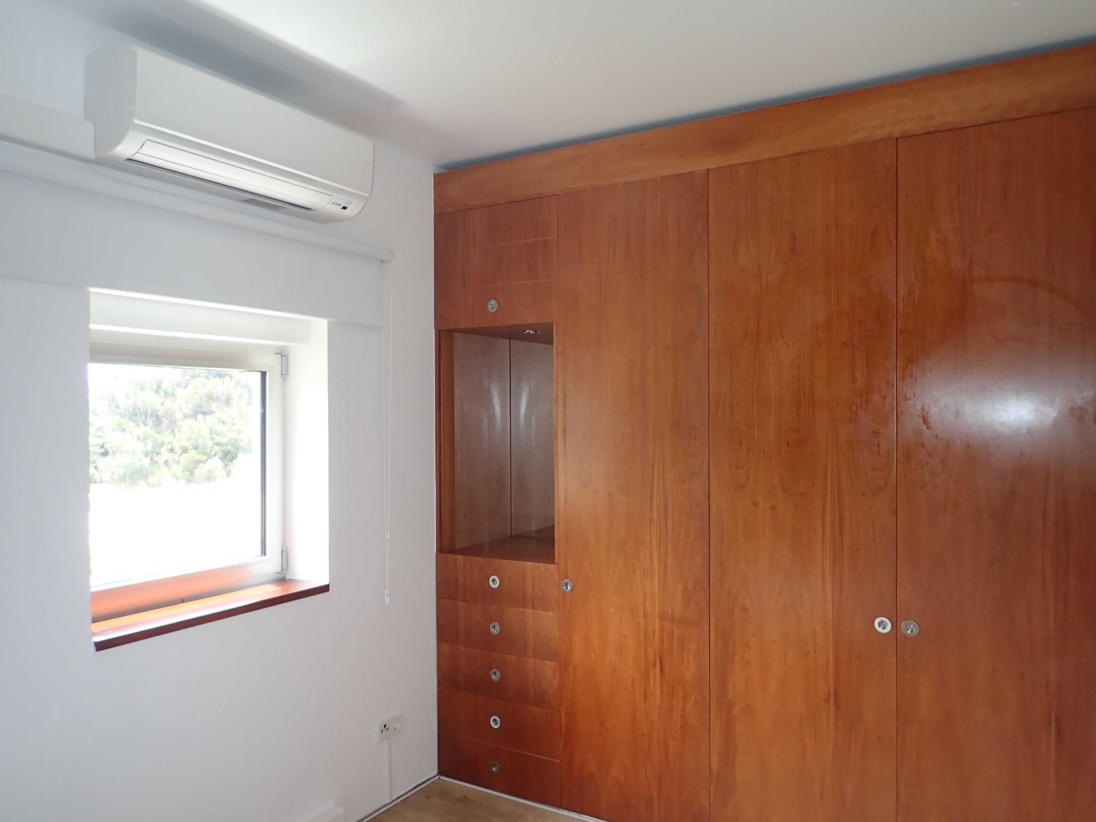 A room with a wooden wardrobe, a window with a view, and an air conditioning unit.