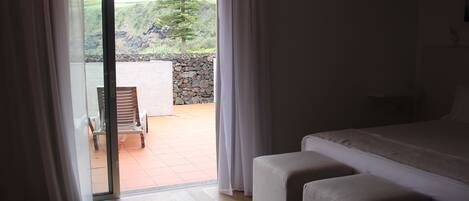 Quarto com cama grande, piso de madeira e vista para um pátio com cadeira e parede de pedra.