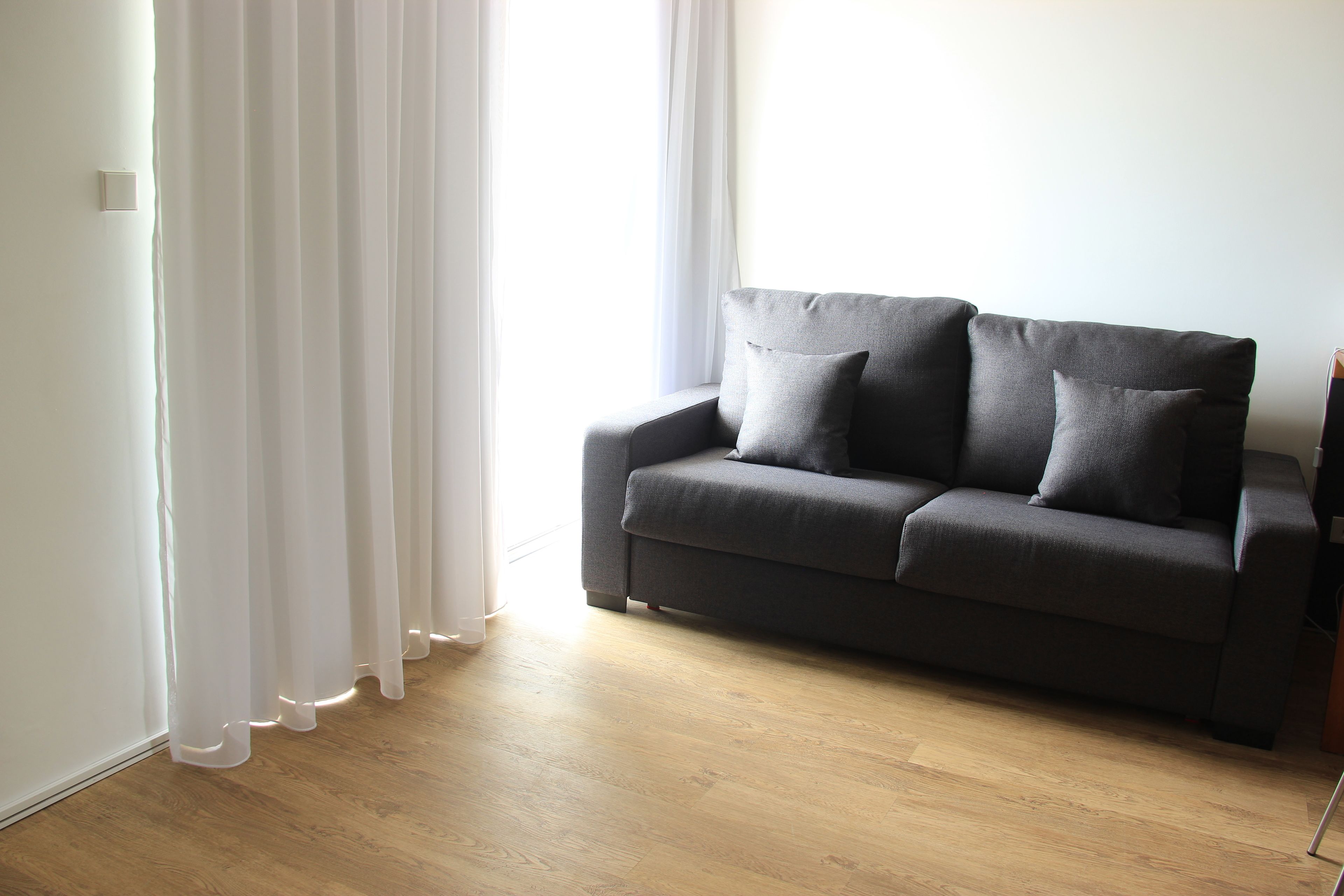 A modern living room with a grey sofa, white curtains, and wooden flooring.