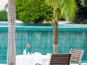 Ein Tisch am Pool mit weißen Tischdecken und Holzstühlen, umgeben von Palmen und einer grünen Hecke.