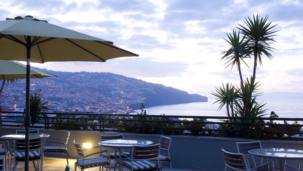 An outdoor seating area with tables and chairs, a large umbrella, and a view of a coastal cityscape. - Madeira Panoramico (Funchal)