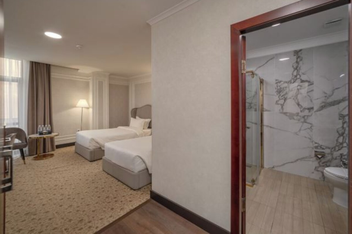 A hotel room with two beds, a sitting area with a table and chairs, a bathroom with a marble shower, and a view of the city.