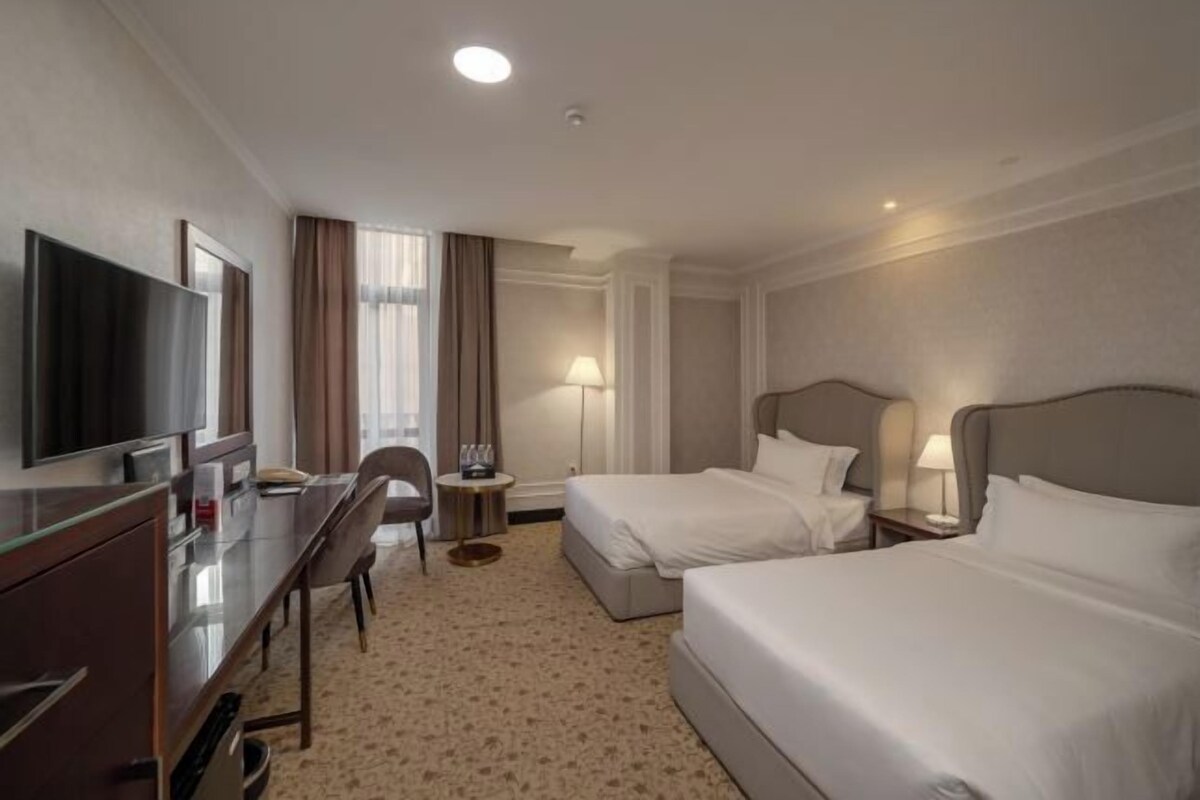 Quarto de hotel com duas camas, uma mesa de escritório com televisão, uma cadeira, um abajur e uma mesinha.