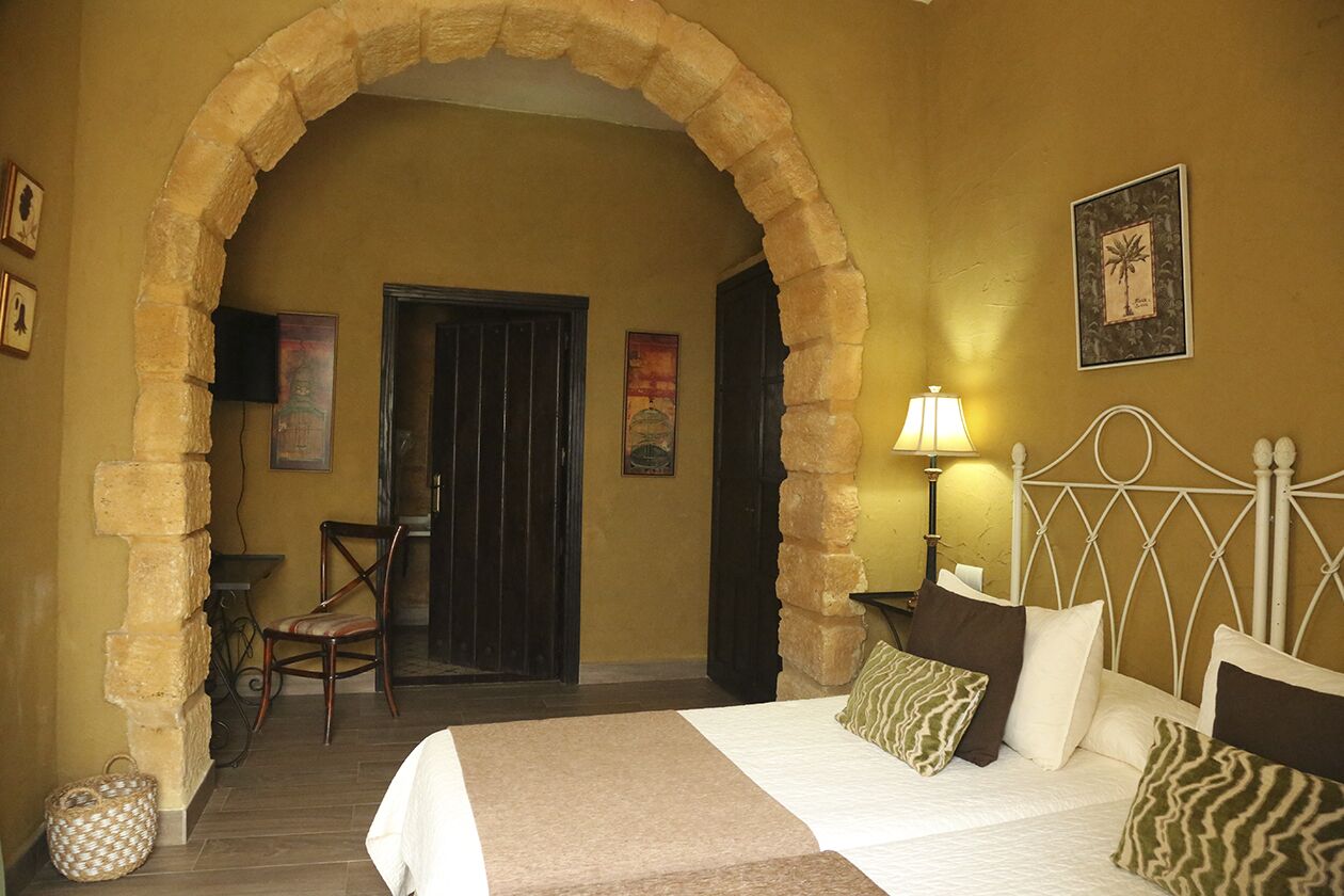 A hotel room with a stone archway, a bed with multiple pillows, a nightstand with a lamp, and a chair.
