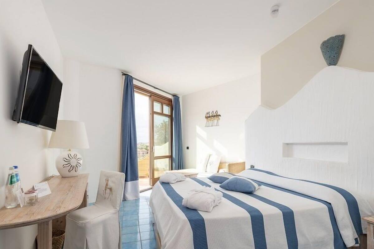 Habitación de hotel con cama, escritorio, silla, televisor y ventana con cortinas.