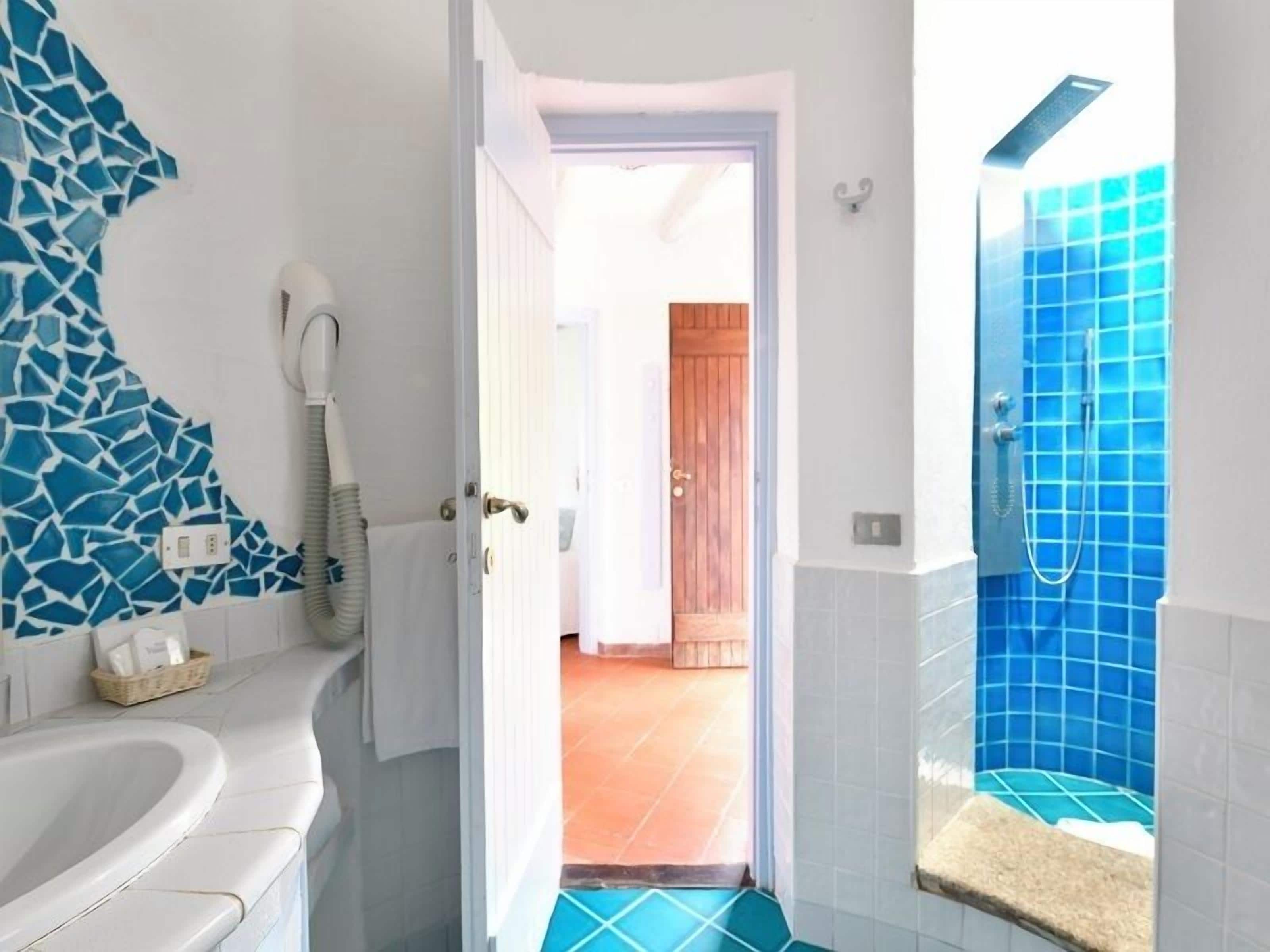 A bathroom with a white sink, a blue mosaic wall, a shower with blue tiles, and a door leading to a room with a tiled floor.
