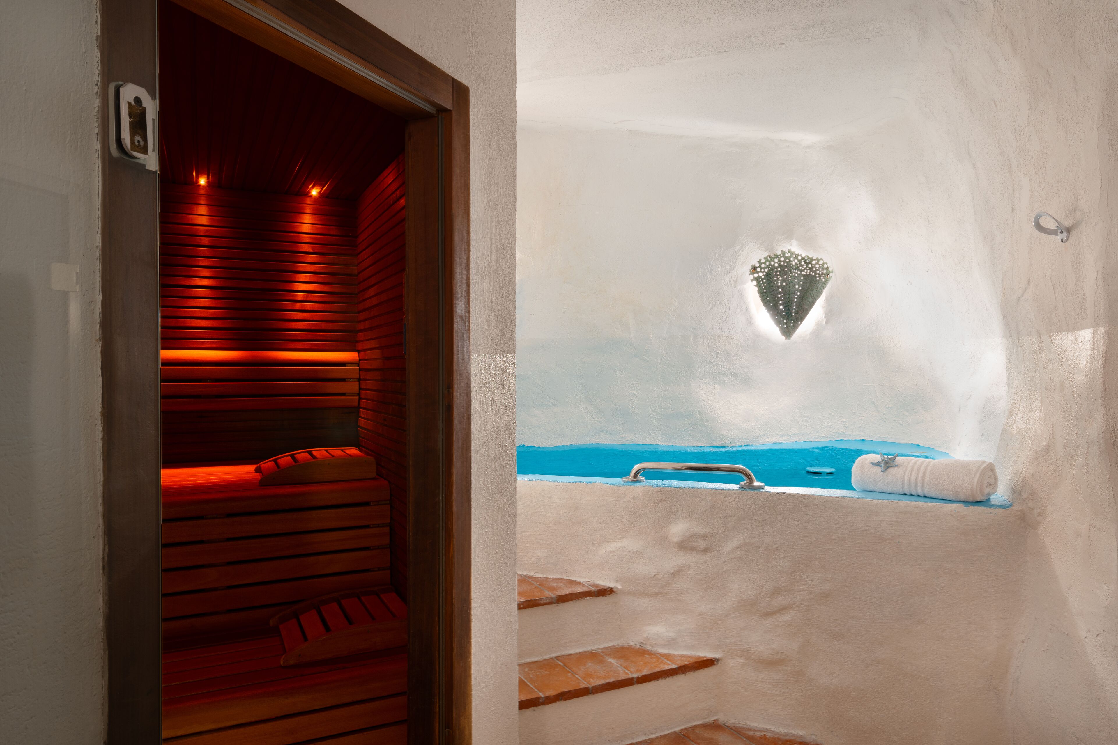Uma sauna com bancos de madeira e chão revestido de azulejos azuis, uma pequena janela com um objeto decorativo, e uma toalha no chão.