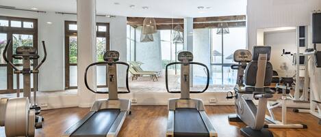 A gym with treadmills, elliptical machines, and weight equipment. Large windows provide natural light and views of the outdoors.