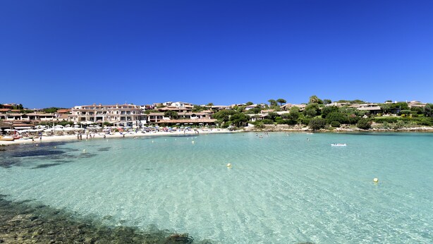 Une zone côtière avec une eau turquoise cristalline, des parasols de plage et des bâtiments en arrière-plan.