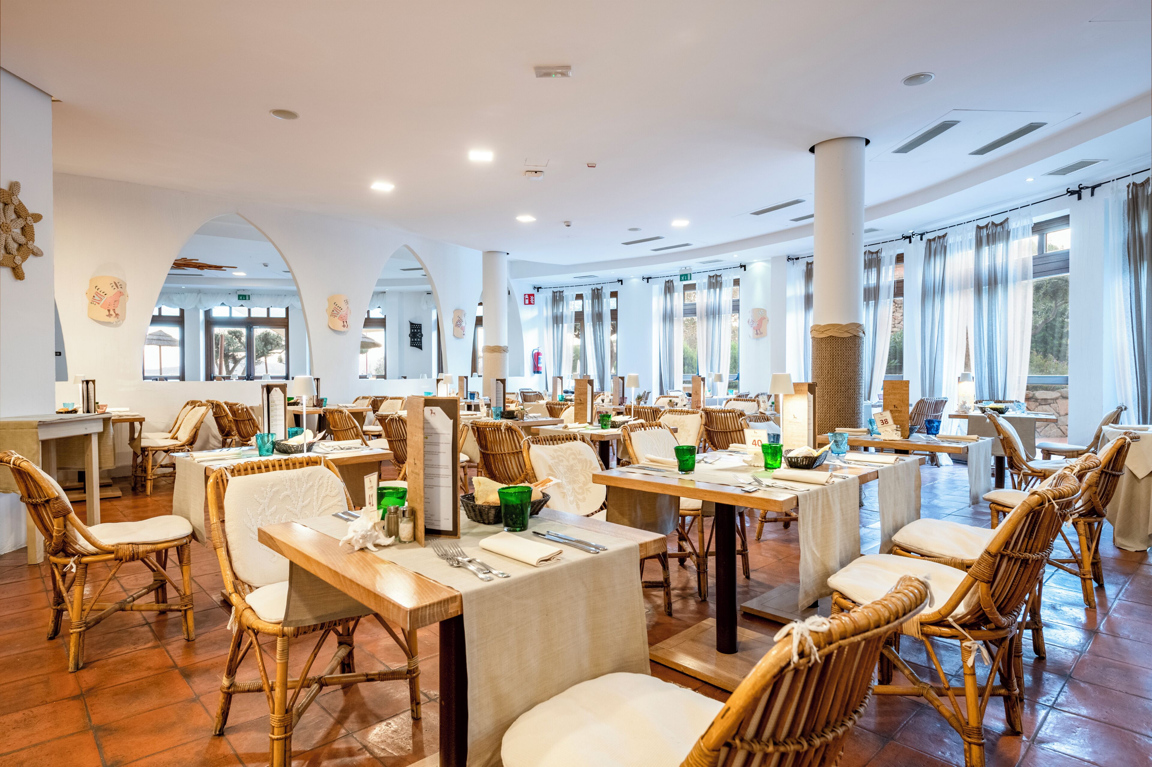 Un restaurante con mesas altas y sillas de mimbre, ventanas amplias y suelo de baldosas.