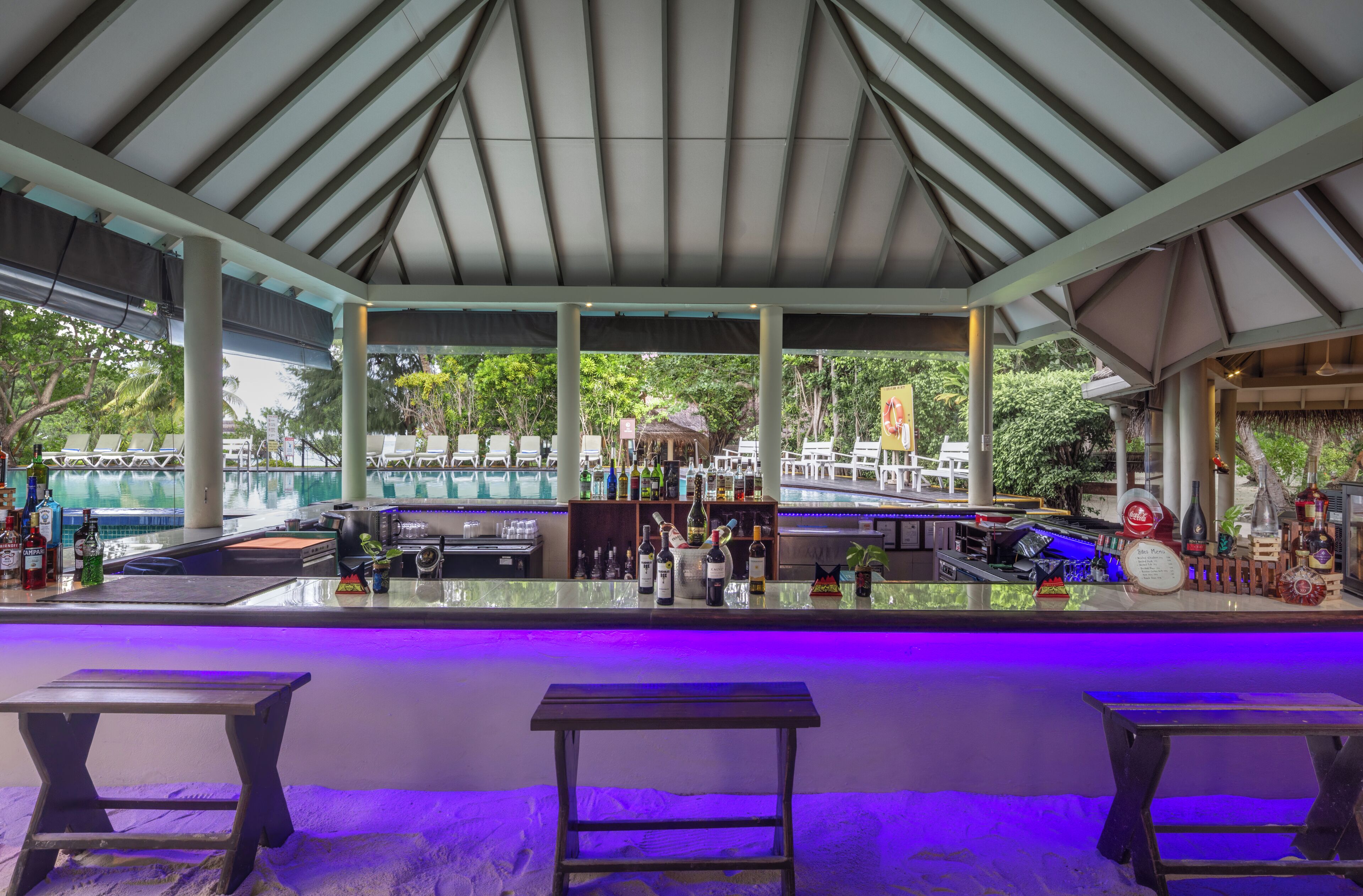 Un espace bar extérieur abrité, avec une piscine en arrière-plan, des tabourets de bar et une variété de bouteilles et de verres disposés sur le comptoir.