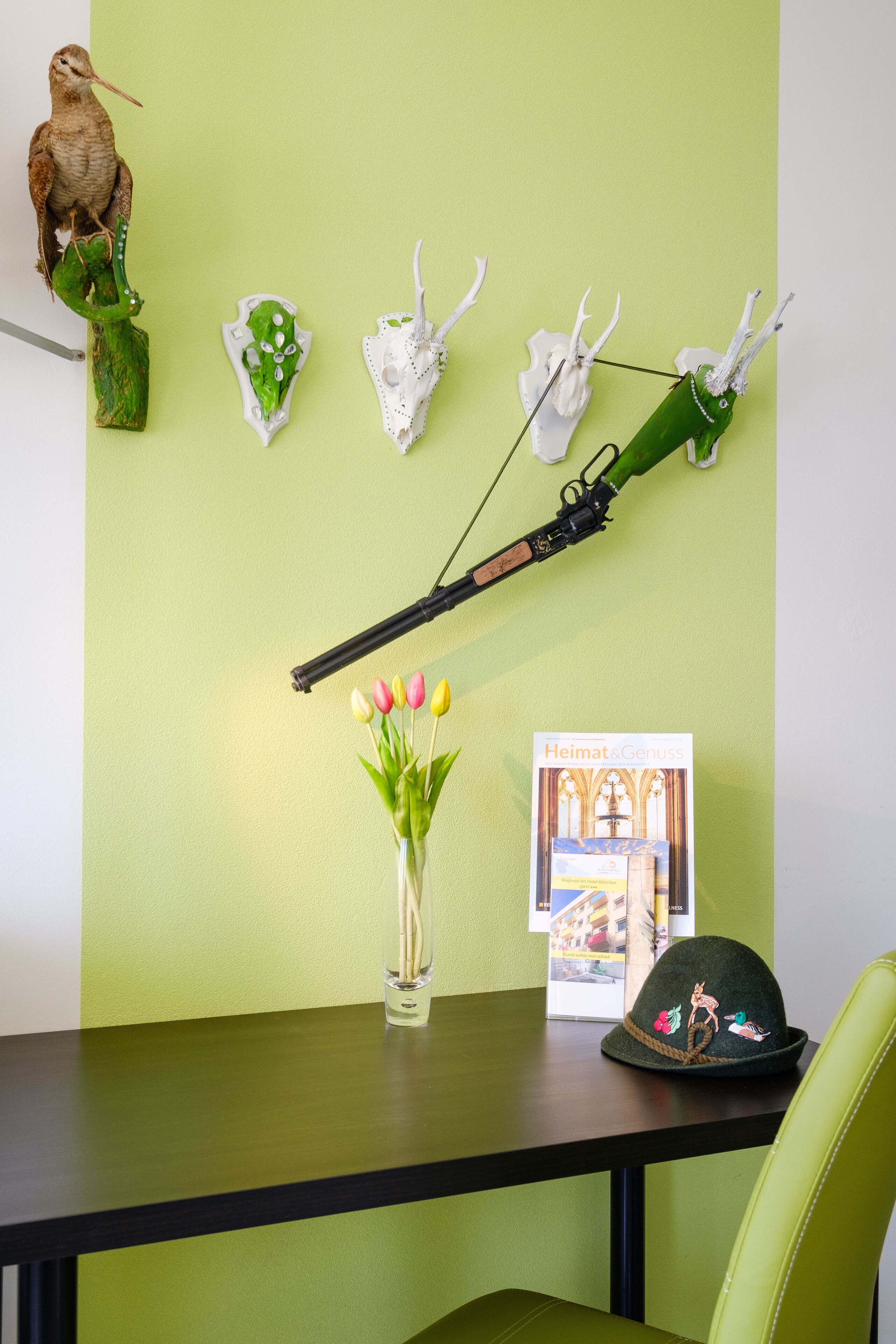 A table with a vase of tulips, a book titled "Heimats Genuss," and a hat with decorative embroidery.