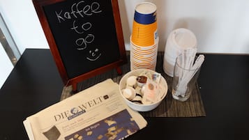 Uma mesa com um jornal, um recipiente com petiscos, uma chávena de café e um letreiro que diz "Kaffee to go".