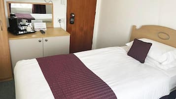 A hotel room with a bed, a wooden desk with a coffee maker, a mirror, and a door with a sign.