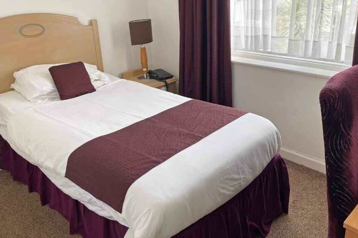 A neatly made bed with a white and maroon bedspread, a wooden headboard, a bedside table with a lamp, a phone, and a chair.