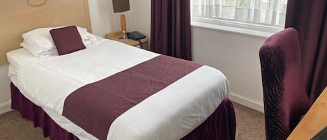 A neatly made bed with a white and maroon bedspread, a wooden headboard, a bedside table with a lamp, a phone, and a chair.