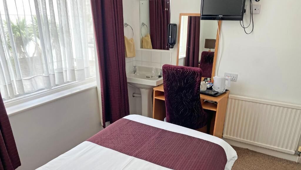Habitación de hotel con cama, escritorio con silla, televisor, espejo, ventana con cortinas y radiador.