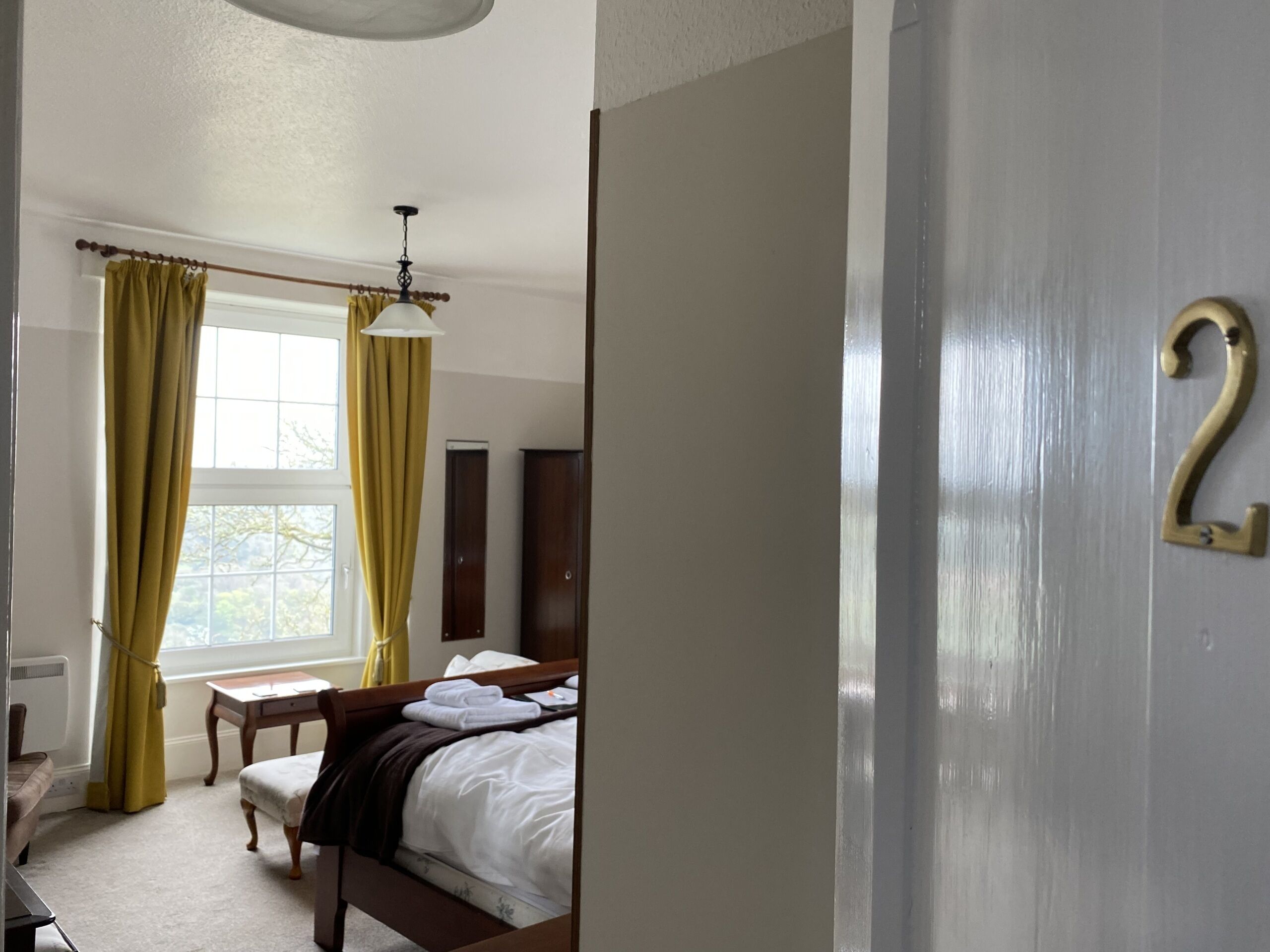 Une chambre à coucher comprenant un lit, une table de chevet, une fenêtre avec des rideaux, un ventilateur de plafond et une porte portant un numéro.