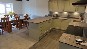 A modern kitchen with wooden chairs around a dining table, a built-in island with a sink, and a view of the outdoors through a window. - Smugglers Rock House (Scarborough)