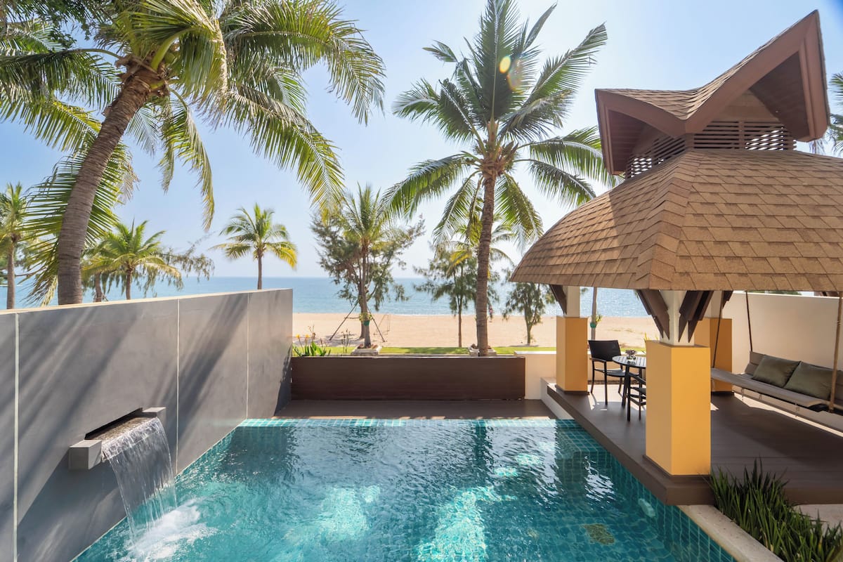 A pool area with a view of the beach, palm trees, and a covered seating area.