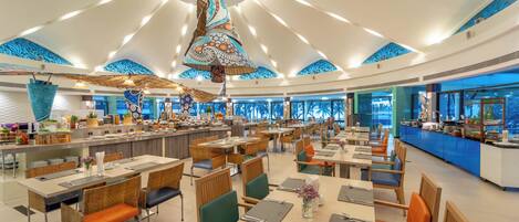 A spacious dining area with high ceilings and a central hanging light fixture. Tables are arranged in rows with chairs, and a counter is visible in the background.