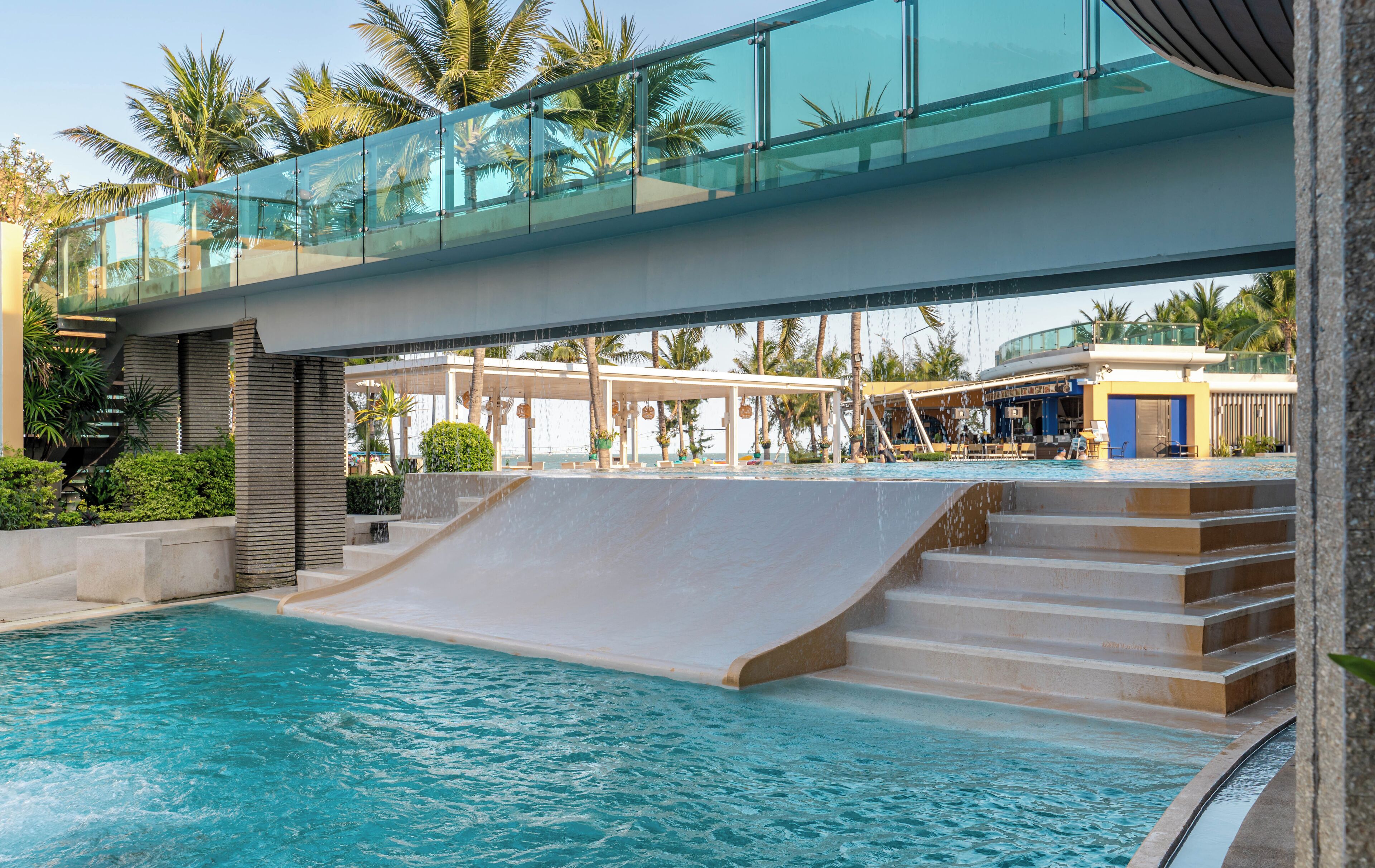 A swimming pool with a glass bridge, palm trees, and a covered seating area.