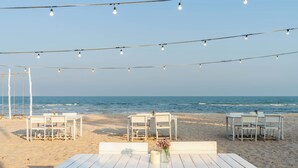 Un espace de restauration aménagé sur la plage, avec des tables et des chaises blanches, des guirlandes lumineuses suspendues au-dessus et une vue sur l’océan.