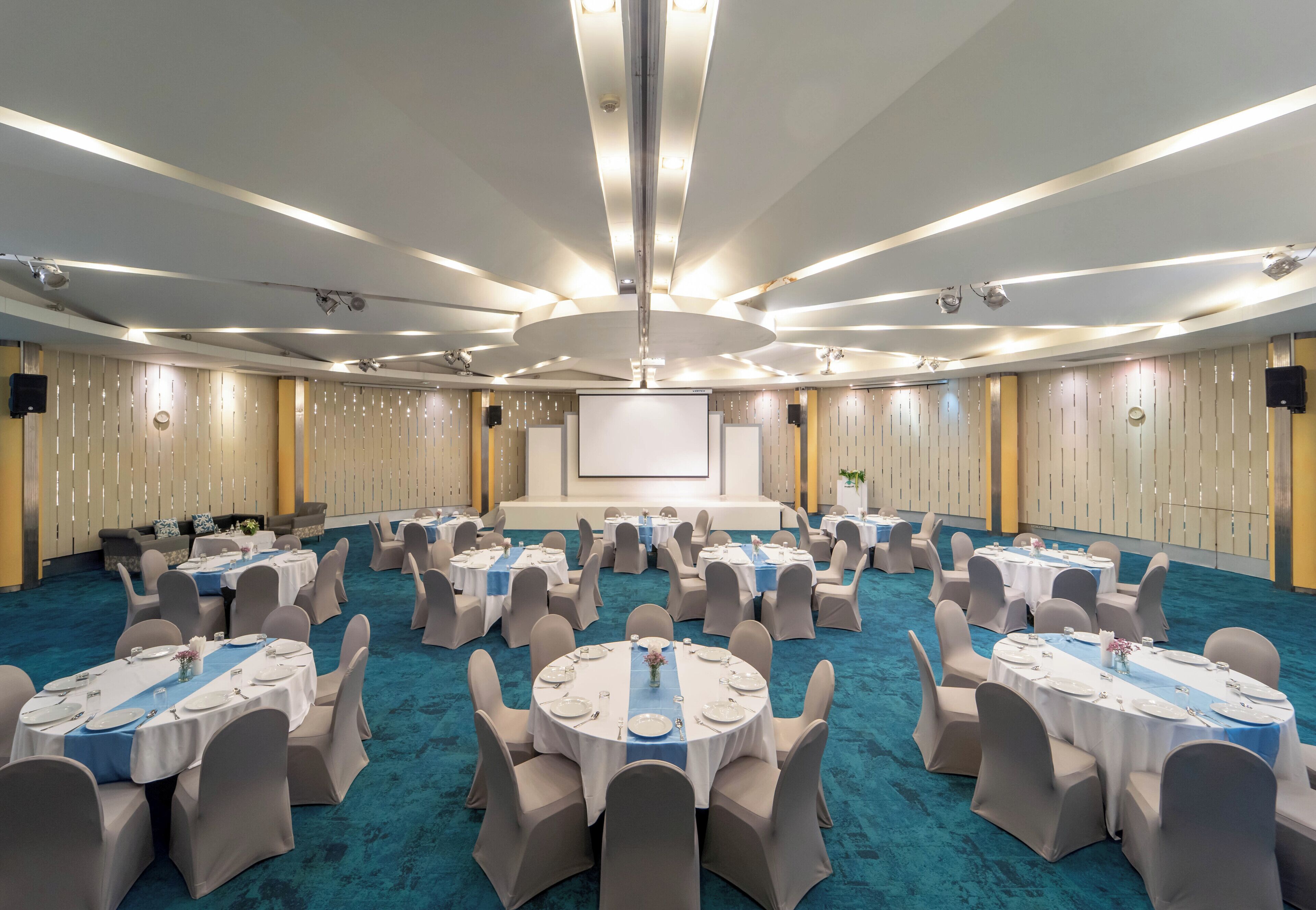 A conference room with multiple round tables, each set with white tablecloths and chairs, arranged in a U-shape around a central stage.