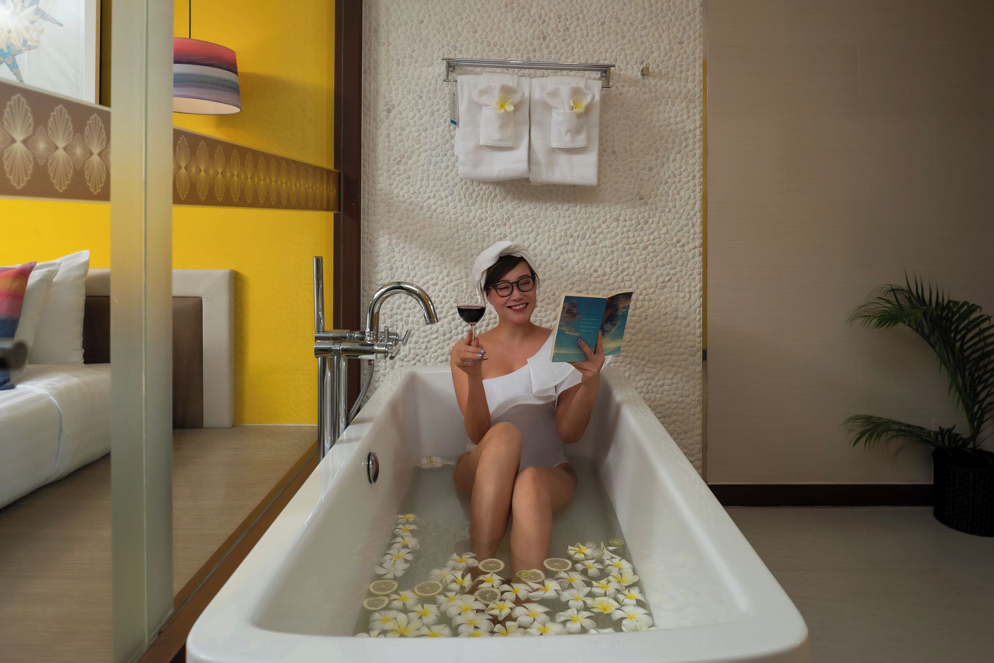 Una persona sentada en una bañera llena de flores, sosteniendo una copa de vino y un libro.