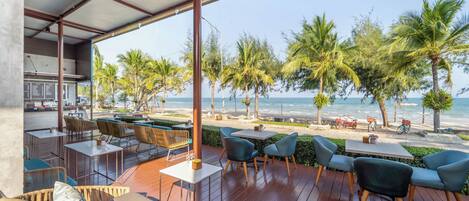 A covered outdoor seating area with tables and chairs, overlooking a beach with palm trees and the ocean.