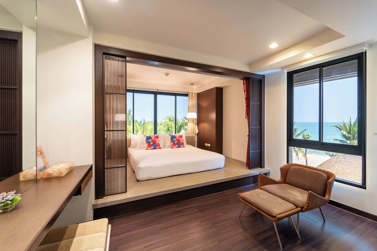 A modern hotel room with a bed, a desk, a chair, and large windows offering a view of the ocean.